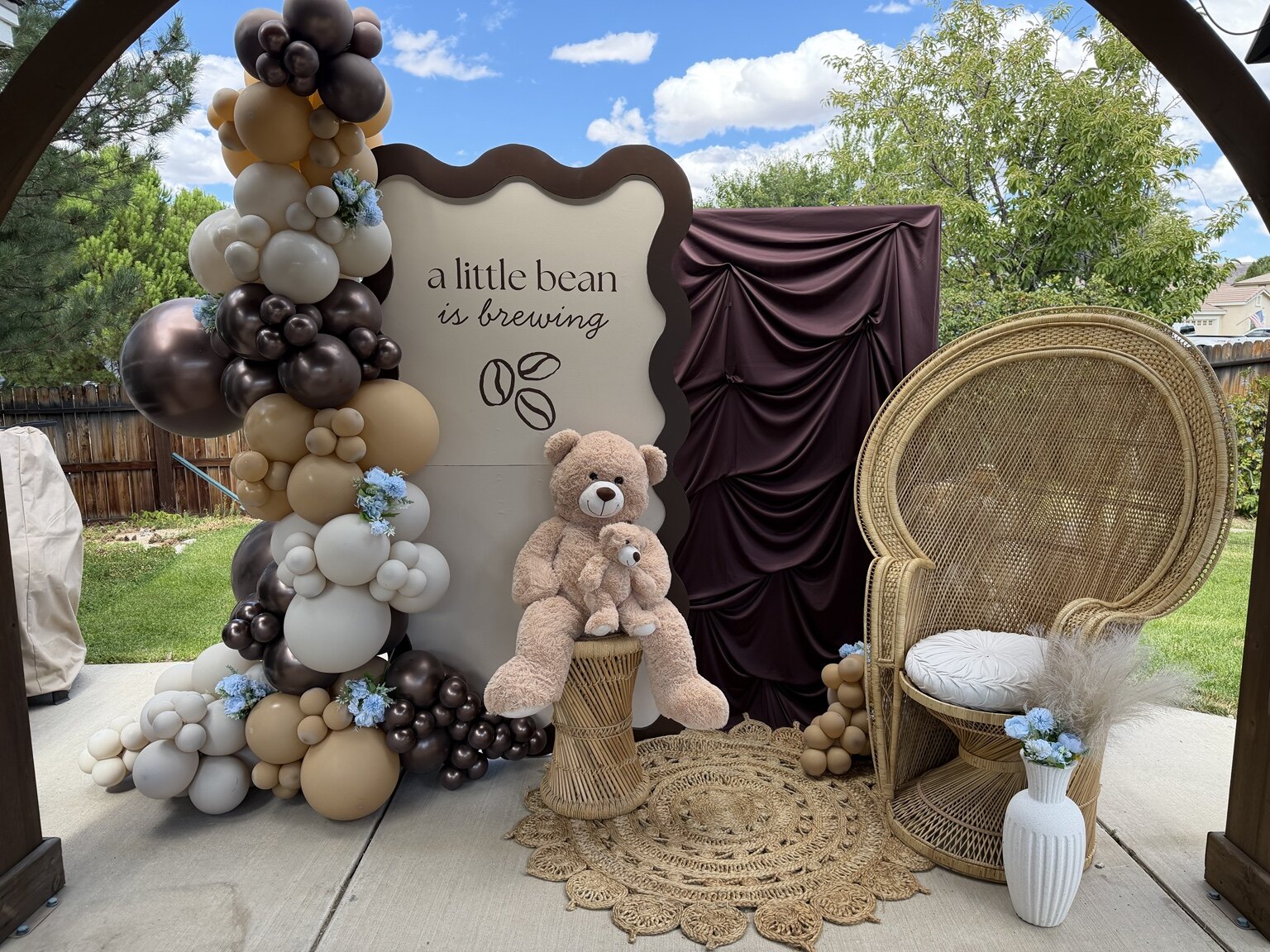 Neutral coffee themed baby shower backdrop with balloons, draping, teddy bears, and peacock chair